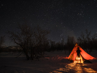 A night photo of arctic landscape with a tent and a silhouette of a person illuminated by a fire in foreground and starry clear sky in the background. (Long exposure time)