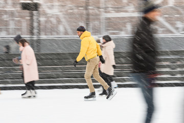 Full length portrait of bearded man in yellow jacket, beige trousers, black hat on ice rink, outdoors in snowy winter day/ Weekend activities outdoor in cold weather/man shooting in motion