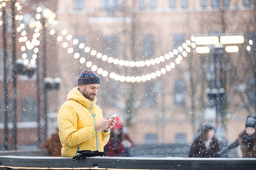 Portrait of joyful bearded man in yellow jacket, black hat standing near fence on ice rink and using smart phone, outdoors at snowy day/Winter time concept/ Weekend activities outdoor in cold weather © DimaBerlin