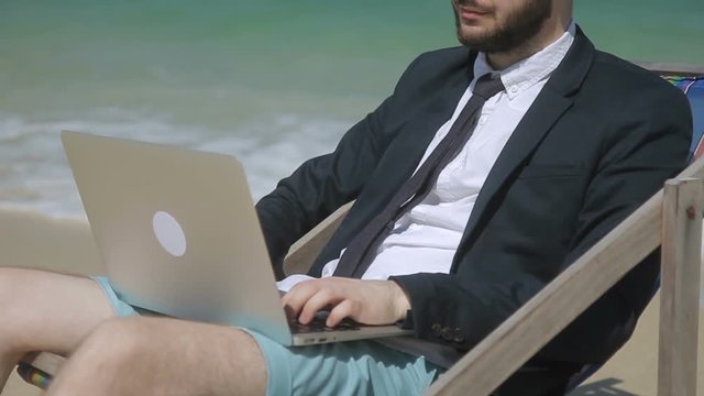 Businessman is typing business email on the beach. Man is sitting on comfortable deckchair with his brand-new laptop on the knees and working. Male professional is in black jacket, tie and blue shorts