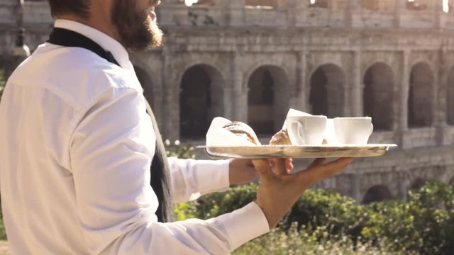 Elegant smiling waiter serving breakfast with breakfast coffee croissant cappuccino on a try outdoor to a bar restaurant table in front of colosseum in rome at sunset