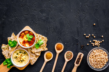 Hummus ready to eat. Bowl with dish among pieces of crispbread and spices on black background top view copy space