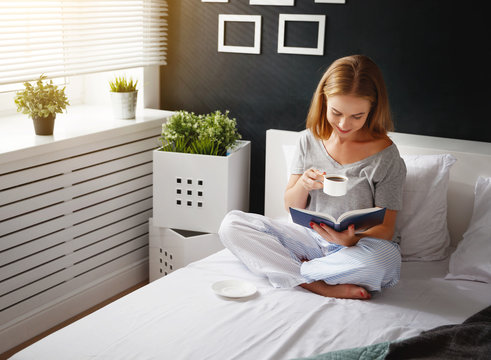 Happy young woman reads  book and drinks coffee in bed