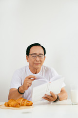 Happy senior couple having croissant and milk breakfast at home, reading book.