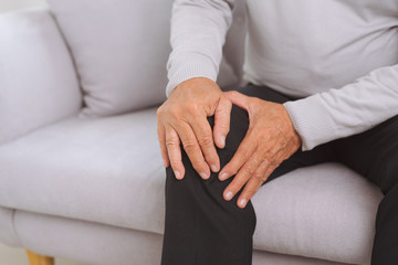 Senior man sitting on a sofa in the living room at home and touching his knee by the pain © makistock