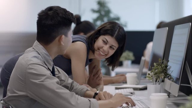 Young Asian male office worker sitting at desk and working on computer while female colleague walking to him and telling something