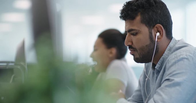 Middle-eastern businessman listening to music with headphones while working on computer in the office, then taking them off and asking colleague about something