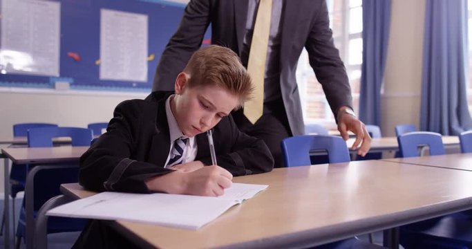 4K Caring teacher checking on the work of young boy on his own in school classroom, could be in detention or child with learning difficulties. Slow motion.