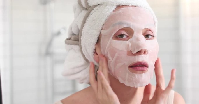 Woman applying cosmetic Japanese mask on her face and looking in the mirror looking at camera in the bathroom