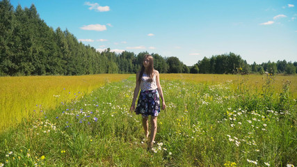Naklejka na ściany i meble Girl walks the field, among flowers and grass. Young girl going away in green field.