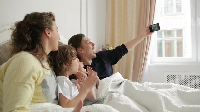 Handsome Man is Taking a Photo by His Smartphone. Positive Family Selfie with Mother, Father and Two Children Lying on the Bed and Wearing Pajamas.