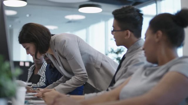 Diverse team of office workers sitting at desk while their female colleague walking with laptop and showing them something on screen