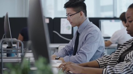 Busy office workers typing on computers in open space office; focus on young Asian clerk working at desk - Powered by Adobe