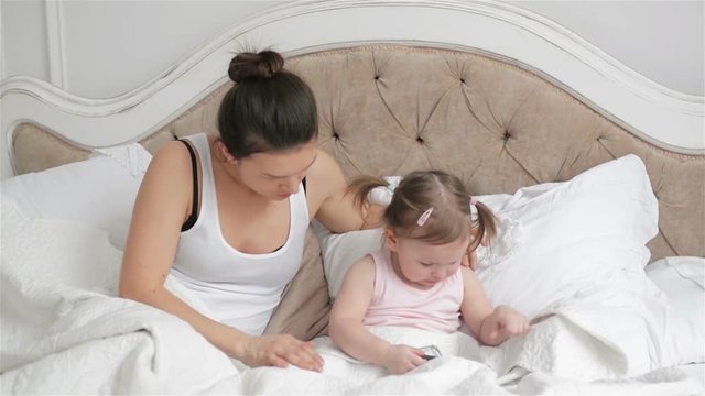 Mother is Trying to Calm Her Daughter in the Bedroom Giving Her Smartphone. Cute Little Girl with Two Ponytails is Crying Sitting on the Bed.