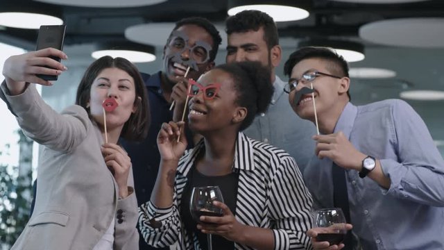 Joyous multi-ethnic team of business men and women holding funny mustache and eyeglasses on sticks, smiling and posing at camera of smartphone while taking selfie at corporate party
