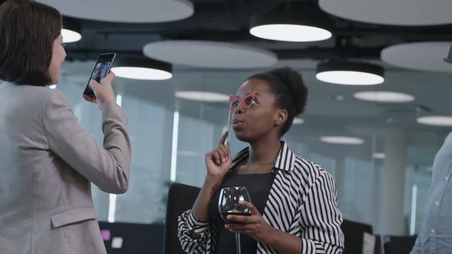 Young African business lady holding paper eyeglasses on stick and posing at camera with glass of wine while female colleague photographing her with smartphone at corporate party