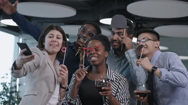 Multi-ethnic team of business men and women holding paper mustache and eyeglasses on sticks, smiling and making funny faces at camera of smartphone while taking selfie together at party in the office