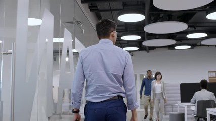 Rear view of businessman going through modern office with stylish hi-tech interior and multi-ethnic employees walking and working at desks