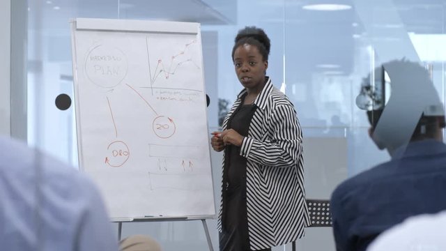 Smart African businesswoman explaining scheme on whiteboard while giving presentation to colleagues in modern meeting room with glass walls