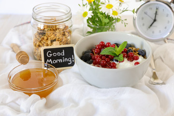 A bowl with yogurt, granola, blueberries and cranberries, granola jar, honey and flowers, alarm clock, good morning and healthy breakfast concept.