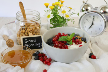 A bowl with yogurt, granola, blueberries and cranberries, granola jar, honey and flowers, alarm clock, good morning and healthy breakfast concept.