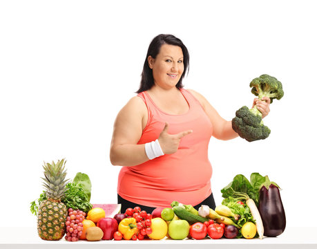 Overweight woman with a broccoli dumbbell pointing behind a table with fruit and vegetables