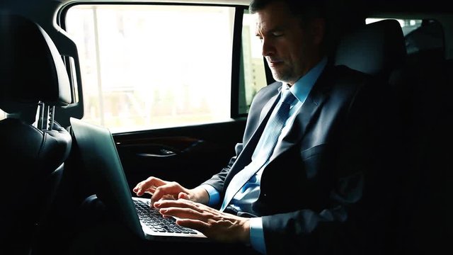 Businessman Riding in Back Seat of Car Working on Laptop.