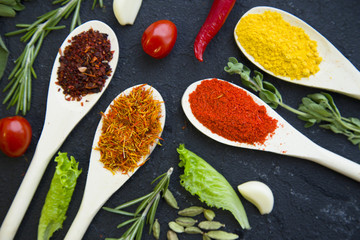 Cooking a hot spicy meal with a set of spices and herbs in wooden spoons, fresh rosemary, lettuce, garlc, and cayenne chili pepper, black concrete background, top view