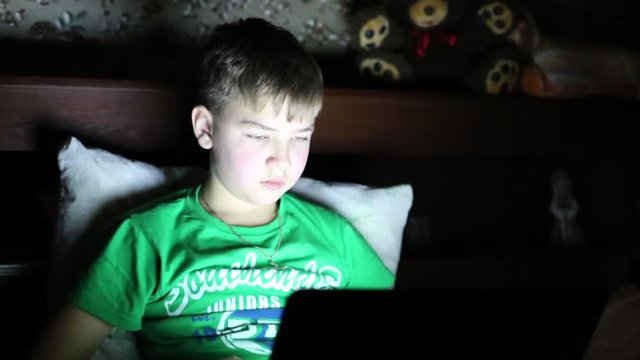 A teenager in a dark room plays computer games. Young boy at night working at computer.