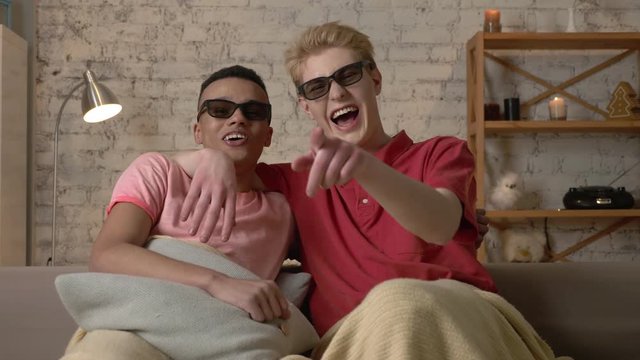 Two gay guys are sitting on a sofa covered in a warm blanket and watching a 3d funny film, they are happy and laughing. LGBT lovers, happy gay family, home cosiness concept. Look at the camera 60 fps