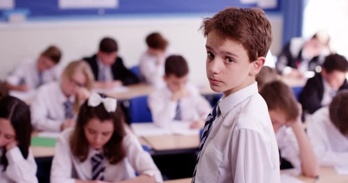 4K Portrait of serious young boy standing in school classroom with his classmates working at their desks. Slow motion.