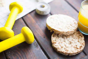 Healthy Breakfast, Sport, Fitness, Diet Concept. Wheat crisp breads, a glass of orange juice, a measure tape and yellow dumbbells on a dark woodeen table