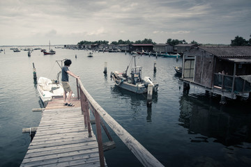 Fototapeta premium Desaturated lake landscape with a boy on a wooden pier watching fishing boats and fishing sheds. Po Delta park lagoon. Veneto, Italy.