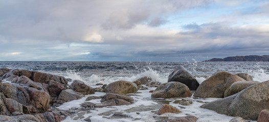 Beautiful seascape of the coast of the Arctic Ocean. Spray, drops of water, white foam and waves. Against the background of a cloudy sky with a mountain on the horizon.
