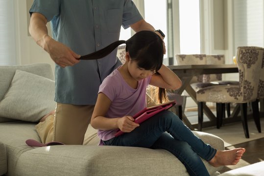 Girl using digital tablet while father combing her hair