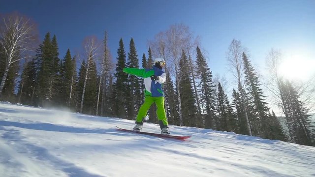 Sportsman in bright green clothes performs a trick on snowboard upon slope of the hill at sunny winter day