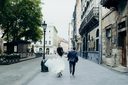 Back view of the glamout gorgeous newlywed couple holding hands while running along the sunny city street.