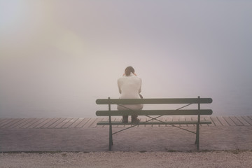 Anonyme junge Frau sitzt betrübt und einsam auf einer Bank im Nebel - obrazy, fototapety, plakaty