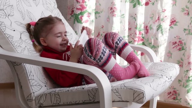 A cute little girl is sitting in armchair near the window playing with digital tablet