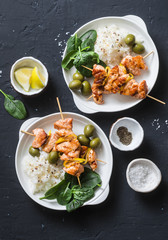 Salmon skewers, olives, spinach, rice - healthy lunch table. Grilled salmon fish skewer and side dish on a dark background, top view