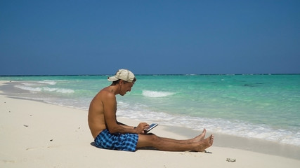 Fototapeta na wymiar Man using electronic tablet on the beach. Man sits on a sandy beach and using a tablet. Tropical beach, blue sky, clouds. Philippines, Travel concept.