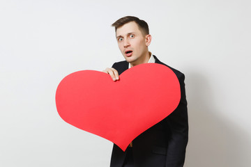 Young stunned shocked upset sad man in suit holding big red heart isolated on white background. Copy space for advertisement. St. Valentine's Day, International Women's Day, birthday, holiday concept.