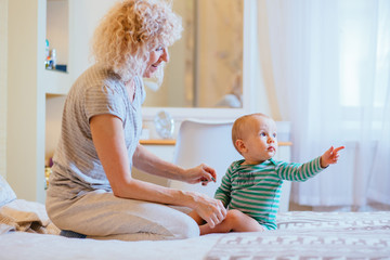Beautiful curly blond grandmother with her infant baby grandson sitting on the bed at cozy bvedroom and having fun together. Real people life concept.