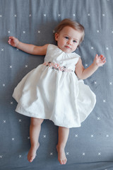 Portrait of cute adorable smiling Caucasian blonde baby girl one year old in white dress lying on grey bed sheet in bedroom, view from top above. Happy childhood lifestyle concept
