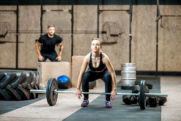Fototapeta na wymiar Young athletic woman lifting up a burbell with man training on the background in the crossfit gym