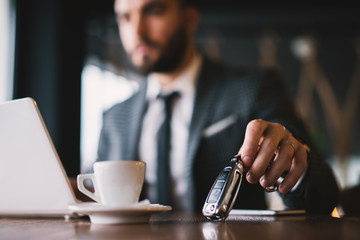 Close view of car keys put on the table next to cup of coffee by rich suited man in the cafe.