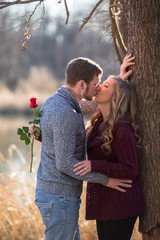 Attractive couple holding hands, kissing,smiling, laughing in love having fun season late winter bare trees near water, lake, red ballon heart shape rub noses