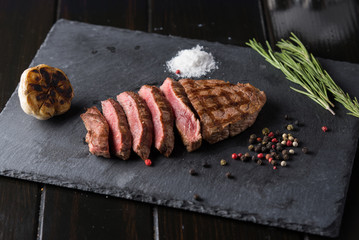 Sliced prime ribeye steak on black stone plate. Medium degree of steak doneness. With rosemary and peppers. Fork and knife.