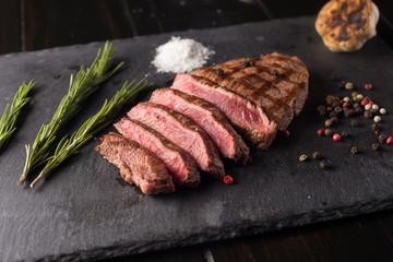 Sliced prime ribeye steak on black stone plate. Medium degree of steak doneness. With rosemary and peppers. Fork and knife.