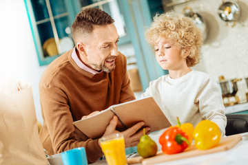 Ready to cook. Pleasant nice joyful man holding a notebook and talking to his son while deciding what to cook